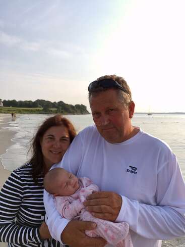 The image depicts a family of three on a beach, with the parents holding a baby, set against a scenic backdrop of a body of water and a grassy area.