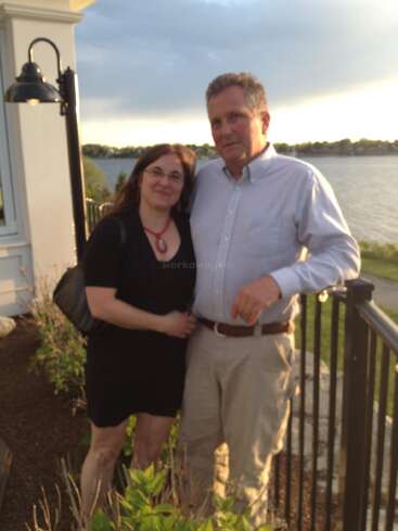 The image depicts a man and woman posing for a photo on a balcony overlooking a body of water, with a cloudy sky in the background.