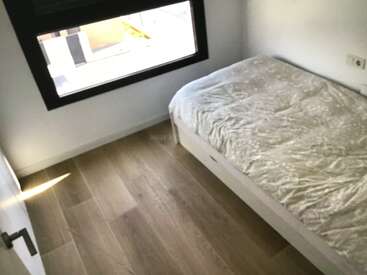 Ein schlichtes Schlafzimmer mit einem Einzelbett, das mit einer hellen Decke bedeckt ist, Holzboden, weißen Wänden, einem großen Fenster und reichlich natürlichem Sonnenlicht, das hereinströmt.