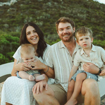 Uma família sorridente de quatro pessoas está sentada ao ar livre. Os pais seguram seus dois filhos pequenos, cercados por árvores verdes e colinas, todos vestindo roupas leves e casuais.