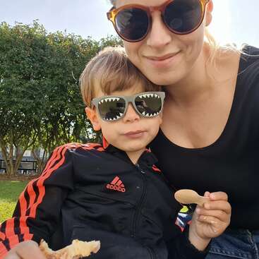 The image shows a woman and a young boy sitting together outdoors, with the boy wearing shark-themed sunglasses and the woman in a black shirt.
