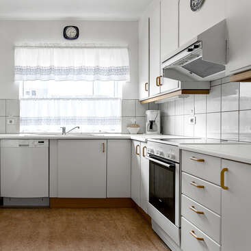 This is a bright, clean kitchen with white cabinets, gold handles, a stove, dishwasher, coffee maker, tiled walls, white curtains, and a wall clock.
