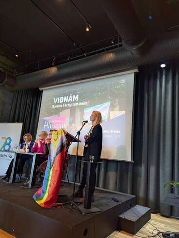 Eine Frau spricht an einem mit einer Regenbogenflagge drapierten Podium. Drei Personen sitzen an einem Tisch. Auf der Leinwand werden isländische Texte und Bilder vom Pride-Event gezeigt.