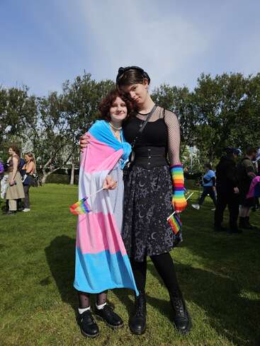 Zwei Menschen stehen zusammen und umarmen sich bei einer Veranstaltung im Park. Sie zeigen ihren LGBTQ+ Stolz mit einer Transgender-Flagge, Regenbogen-Armbändern und Fahnen. Die Sonne scheint, Menschen versammeln sich.