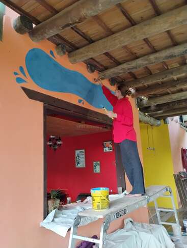 Uma mulher está em uma escada, pintando um desenho azul em uma parede de uma sala com paredes vibrantes e vigas de madeira expostas.