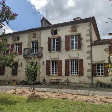 L'image représente une grande maison en pierre à deux étages avec des volets bruns et une allée en gravier, sur fond de ciel nuageux.