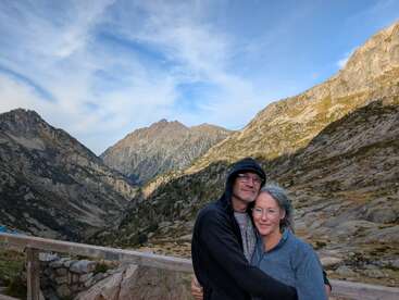 A couple embraces warmly, standing on a wooden terrace amidst rugged mountain scenery. The sky is partly cloudy, and the serene atmosphere enhances their affectionate moment.