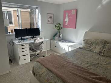 Esta imagem mostra um quarto bem iluminado com uma mesa de trabalho, dois monitores, móveis brancos, uma cama aconchegante, decoração em tons pastéis, obras de arte e uma grande janela.