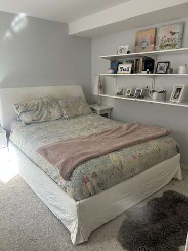 Um quarto aconchegante com uma cama bem arrumada, roupa de cama floral, uma manta lilás, prateleiras de parede com livros, obras de arte, fotos emolduradas, plantas e decoração pessoal. Iluminação suave.