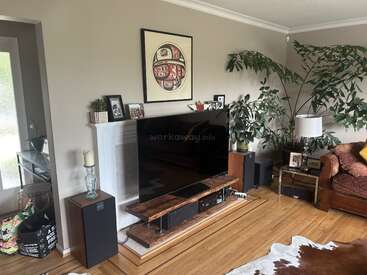 A aconchegante sala de estar possui uma TV grande, alto-falantes, móveis de madeira, obras de arte decorativas, plantas exuberantes, um sofá marrom, uma luminária e um tapete de couro.