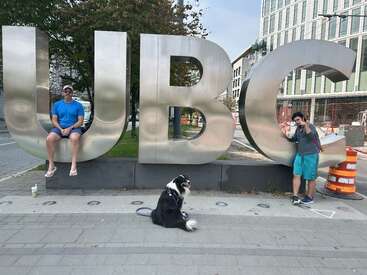 Dois homens e um cachorro preto e branco posam em frente a grandes letras metálicas "UBC"; um senta-se no "U" e o outro fica perto do "C".