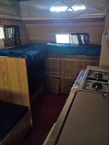 Esta imagen muestra un acogedor interior de caravana con paneles de madera, una cama revestida de azul, pequeñas ventanas, cortinas estampadas, armarios y una compacta zona de fogones.
