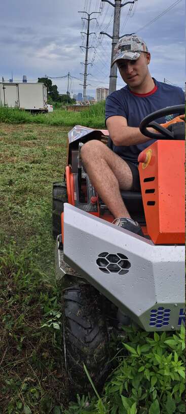 A man in a newspaper-pattern cap drives an orange ride-on mower through grassy terrain. Power lines, green plants, and a cloudy sky set the background.