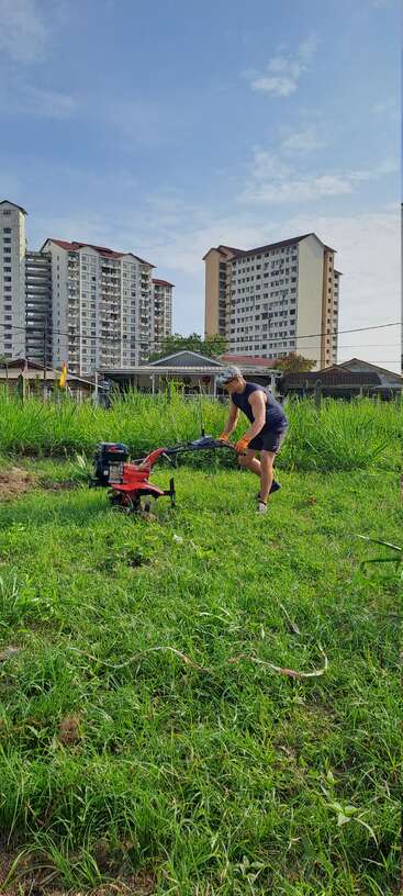 A man in athletic wear operates a red tiller on a grassy field. Tall apartments and blue sky form the urban background, mixing city and nature.