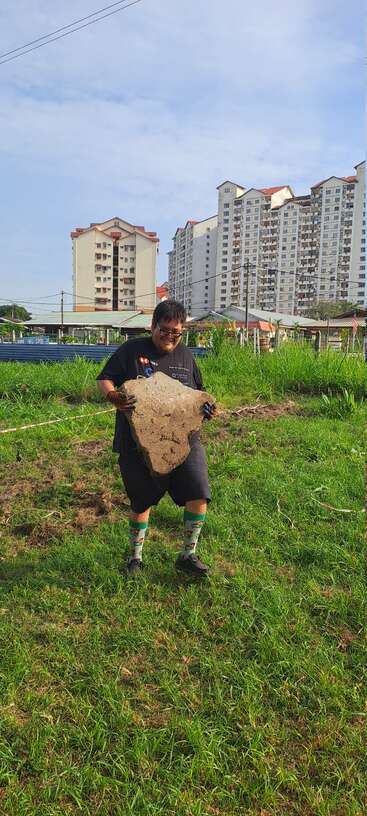 A man in black clothing and quirky socks is lifting a large rock in a grassy field, with tall residential buildings visible in the background.