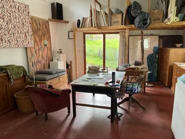 Das Bild zeigt ein unordentliches Zimmer mit einem Schreibtisch, einem Stuhl und verschiedenen Kunstsachen, mit einem Fenster, das einen Blick auf eine Wiese draußen bietet.