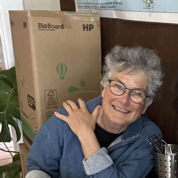 Das Bild zeigt eine Frau mit kurzen grauen Haaren, die eine blaue Jacke trägt und mit der Hand auf der Schulter vor einem Pappkarton steht.