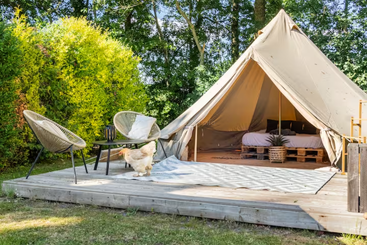 Une tente de glamping confortable se dresse sur une terrasse en bois, entourée de verdure. À l'intérieur se trouve un lit confortable. Deux chaises, une table et un poulet complètent le décor.