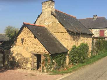The image depicts a charming stone cottage with a sloping roof and chimney, nestled among other buildings in a quaint village setting on a sunny day.
