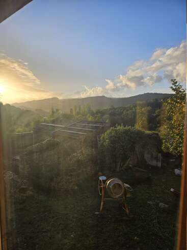 Eine schöne ländliche Landschaft bei Sonnenuntergang, mit sanften Hügeln, üppigem Grün, einer alten Steinmauer, Dächern und einem Zementmischer im Vordergrund, durch Glas betrachtet.