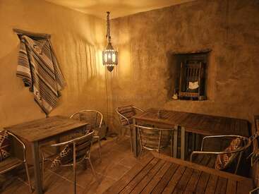 Warm beleuchteter, rustikaler Raum mit hellbraunen, strukturierten Wänden, Holztischen, Metallstühlen, gemusterten Kissen, gewebter Decke an der Wand, hängender Laterne und einer kleinen Fensternische.