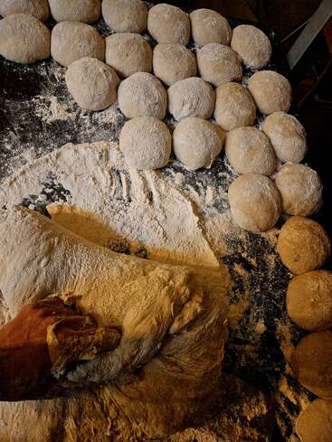 Eine Hand knetet ein großes Stück Teig auf einer bemehlten Fläche, das von vielen runden Teigkugeln umgeben ist, die zum Backen oder Kochen bereit sind.