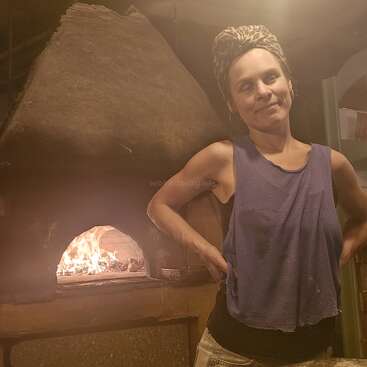 Eine Frau steht selbstbewusst vor einem steinernen Holzofen, in dem ein Feuer brennt. Sie trägt ein Kopftuch und ein ärmelloses Oberteil in einer gemütlichen Umgebung.