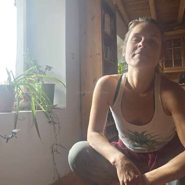 A woman sits indoors near a sunlit window with potted plants. She wears a white tank top, looking relaxed, surrounded by wooden furniture and bookshelves.