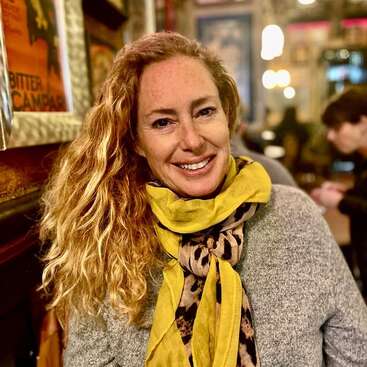 Eine lächelnde Frau mit lockigem rotem Haar und Sommersprossen hat einen gelben Schal mit Leopardenmuster und einen grauen Pullover an und sitzt drinnen in einem gemütlichen Café mit warmer Beleuchtung.