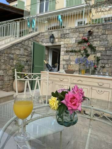 La imagen muestra un entorno sereno al aire libre, con una mesa de cristal con un vaso de zumo de naranja y un jarrón de flores, junto a un muro de piedra y un balcón.