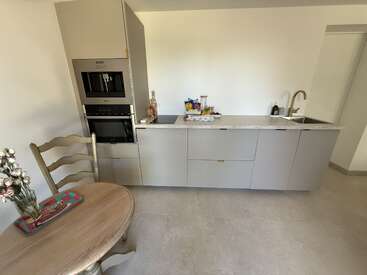 En esta imagen se ve una cocina moderna y minimalista con armarios de color gris claro, electrodomésticos empotrados, una encimera con diversos objetos, un fregadero y una pequeña mesa redonda de madera.