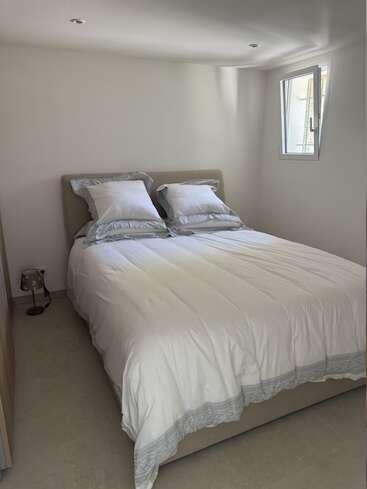 En esta imagen se ve una cama doble perfectamente hecha, con sábanas blancas y ribetes decorativos grises, dos almohadas, una lámpara pequeña y una ventana por la que entra la luz del sol.