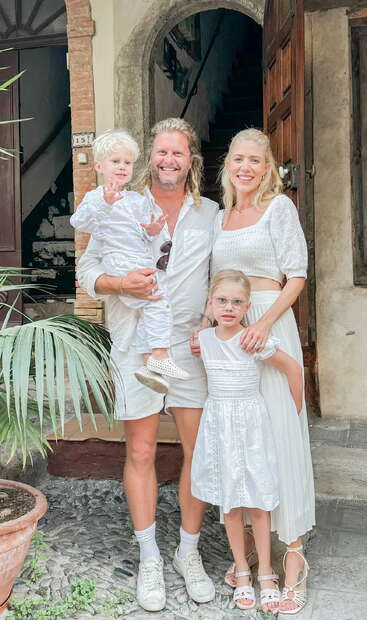La imagen muestra a una familia de cuatro miembros en el exterior de un edificio, con los padres sujetando a sus dos hijos, todos vestidos de blanco, posando para una foto.