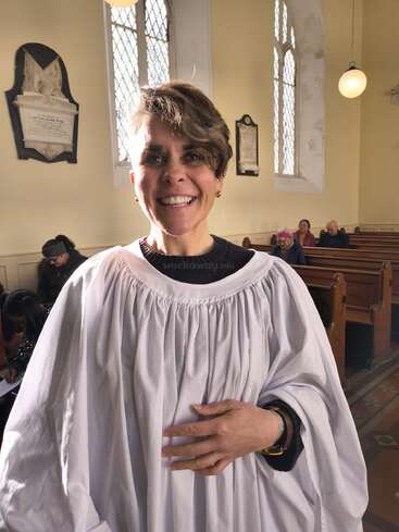 Une femme souriante, vêtue d'une robe de choriste blanche, se tient debout dans une église baignée de lumière, avec en arrière-plan des vitraux et des personnes assises. L'atmosphère est chaleureuse.