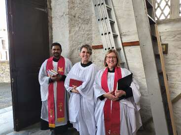 Trois membres du clergé, vêtus de robes blanches et d'étole rouge, se tiennent debout, souriants, à l'intérieur d'une église, un livre à la main, avec un mur de pierre et une échelle en arrière-plan.