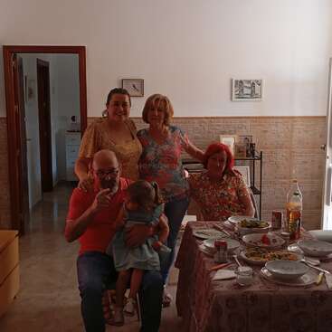 Five people, including a child, gather cheerfully around a dining table with food and drinks. The room is warmly lit, evoking a cozy family atmosphere.