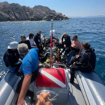 A group of divers sits on a boat, surrounded by scuba gear, heading out to sea with rocky islands in the background under a clear sky.