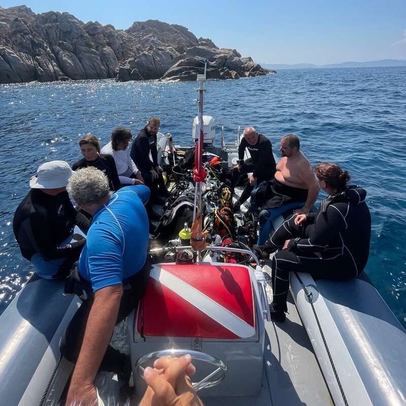 A group of divers sits on a boat, surrounded by scuba gear, heading out to sea with rocky islands in the background under a clear sky.