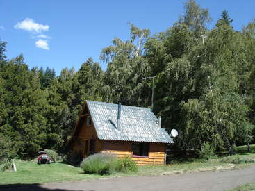 Das Bild zeigt eine kleine, hölzerne Hütte mit grauem Dach, die in einem Waldgebiet liegt, mit einer Satellitenschüssel und einem roten Fahrzeug, das auf der Wiese parkt.