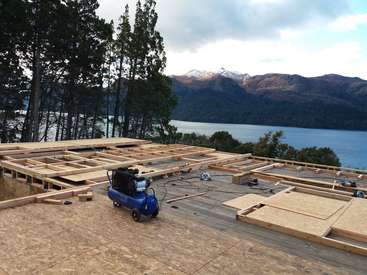 Das Bild zeigt eine Baustelle mit Holzrahmen und Sperrholz auf einem Deck mit Blick auf einen See, umgeben von Bäumen und Bergen im Hintergrund.