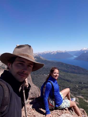 Das Bild zeigt einen Mann und eine Frau, die auf einem Felsvorsprung sitzen, mit einem ruhigen See und Bergen im Hintergrund unter einem klaren blauen Himmel.