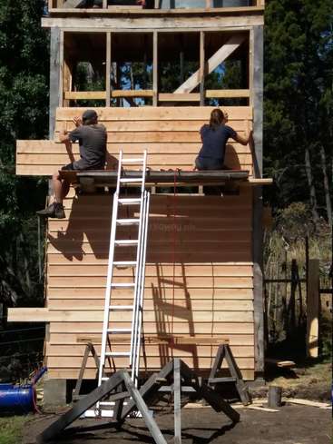 Zwei Menschen bauen eine Holzkonstruktion. Sie sitzen auf der obersten Ebene und lehnen eine Leiter daran, umgeben von Bäumen und einem sonnigen Tag.