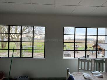 This image shows a cozy room with large windows overlooking a peaceful garden. Outside, there are trees, tables, chairs, a dog house, and dogs.