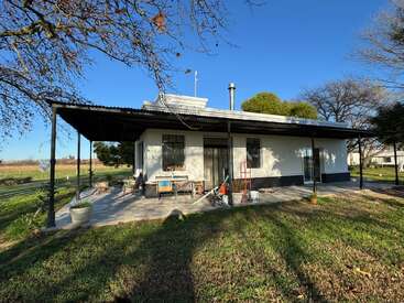 A single-story house with a large covered patio, surrounded by green grass and trees, featuring outdoor furniture, garden tools, and bright daylight under a clear sky.