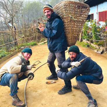 Tres hombres posan para una foto, acuclillados en un camino de tierra, vestidos con ropa de invierno, con una gran cesta de mimbre a la espalda del hombre del centro.