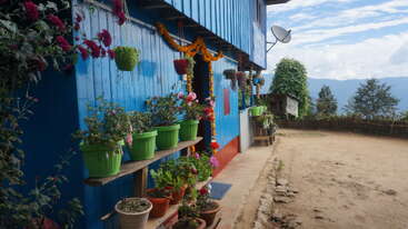 Ein leuchtend blaues Haus mit bunten Topfpflanzen und Girlanden steht auf einem Hügel, und unter dem teilweise bewölkten Himmel sieht man Berge und Bäume.
