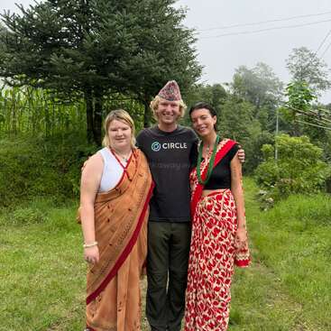 Three people are standing outdoors, smiling. Two women wear traditional South Asian attire, while the man in the middle wears a t-shirt, pants, and a Nepali topi.