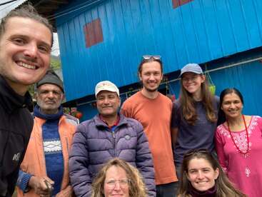 Um grupo animado de oito pessoas está reunido em frente a um prédio azul brilhante, sorrindo para a câmera. Alguns estão bem agasalhados, e o clima é de simpatia.
