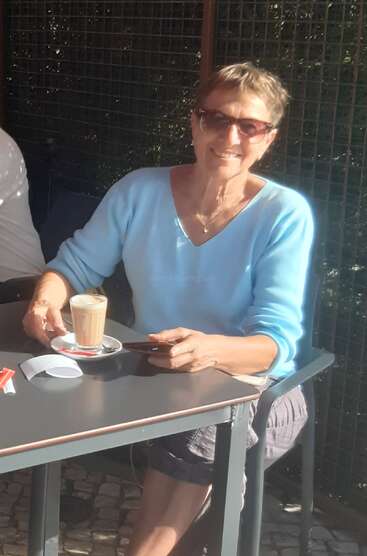 A woman wearing sunglasses and a light blue sweater sits at an outdoor café table, smiling, holding a phone, and enjoying a coffee in the sunshine.