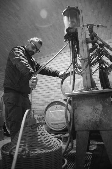 A man in glasses operates industrial equipment in a rustic, brick-walled room, possibly a winery or brewery, surrounded by barrels, pipes, and woven baskets.